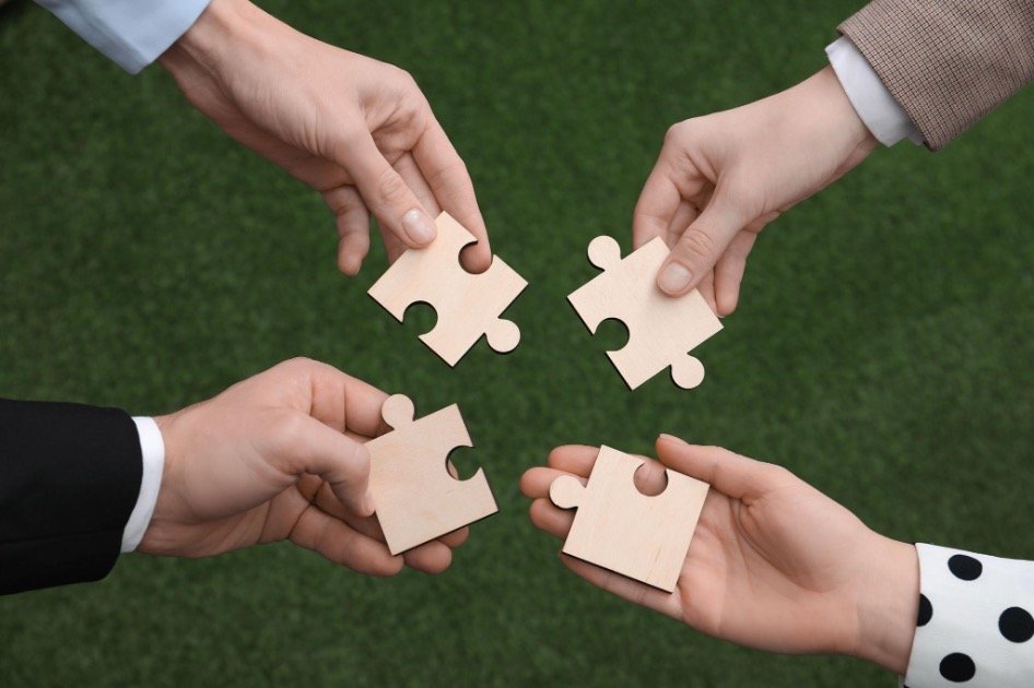 Four hands of different professionals holding wooden puzzle pieces over a green surface, symbolising how different kinds of businesses fit together.