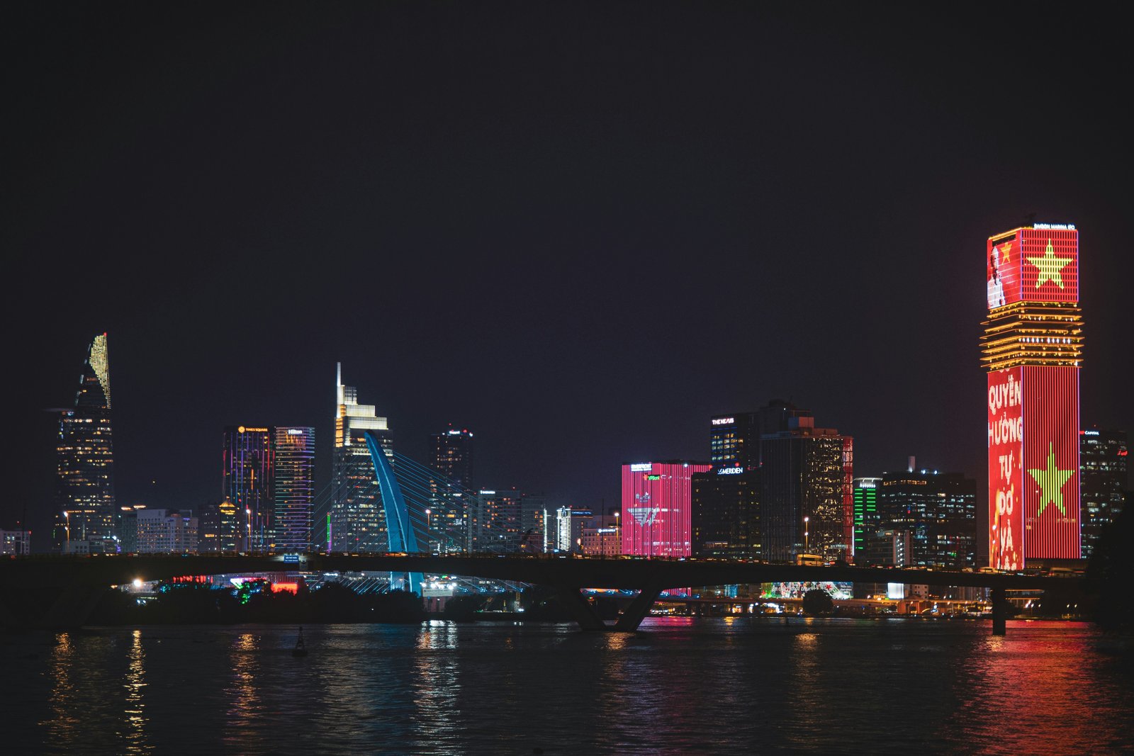 Ho Chi Minh City night skyline reflected on the Saigon river, with a high-rise lit up in the colours and star of the Vietnamese flag, evoking corporate presence and operations in Vietnam.