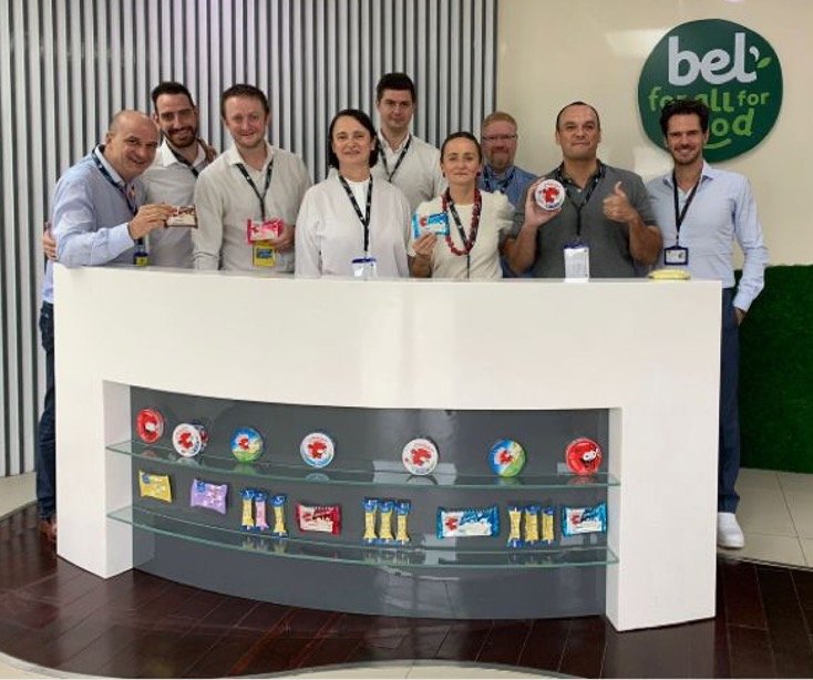 Group photo of nine people at the Bel Vietnam factory reception, with a Bel “For all, for good” sign in the background and Kiri, La Vache qui rit and other Bel Group products displayed on the counter in front.